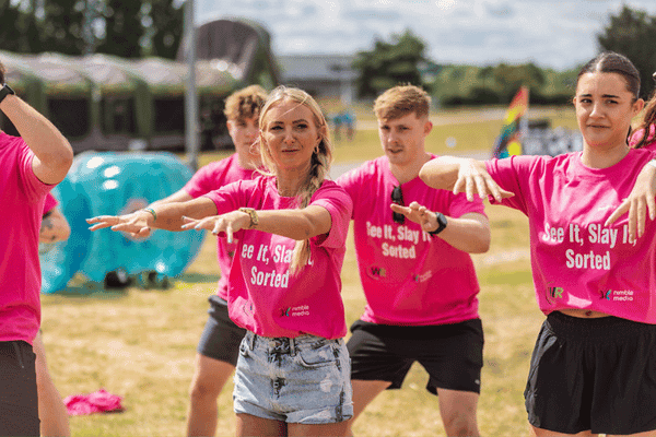 Outdoor team-building event with participants wearing matching blue shirts, taking part in inflatable obstacle courses and games, smiling and cheering, with the words 'ENTER A TEAM' in large bold white and green text across the image.