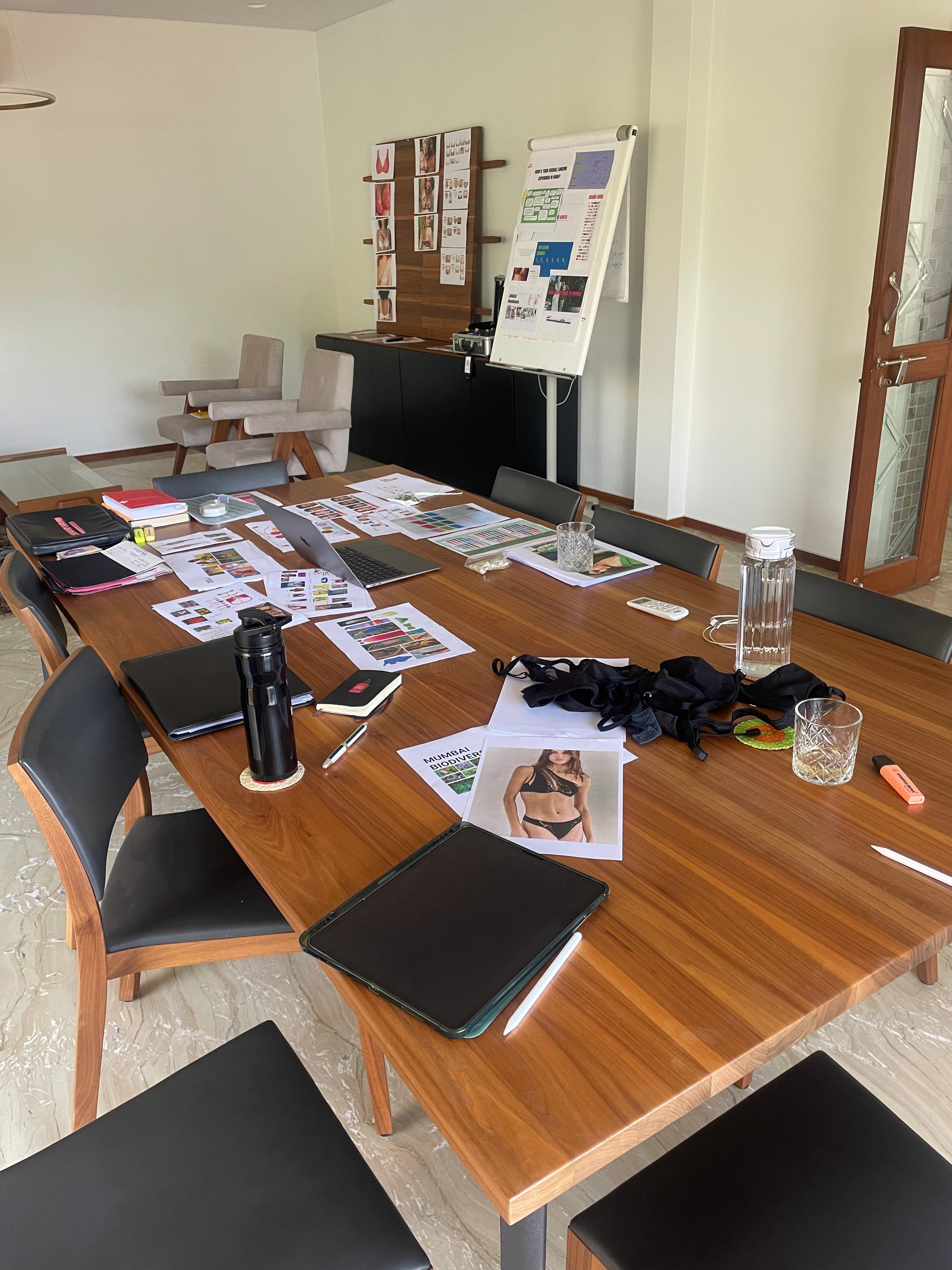 Wooden table with lingerie samples, moodboards, and drinks set up for a creative work session.