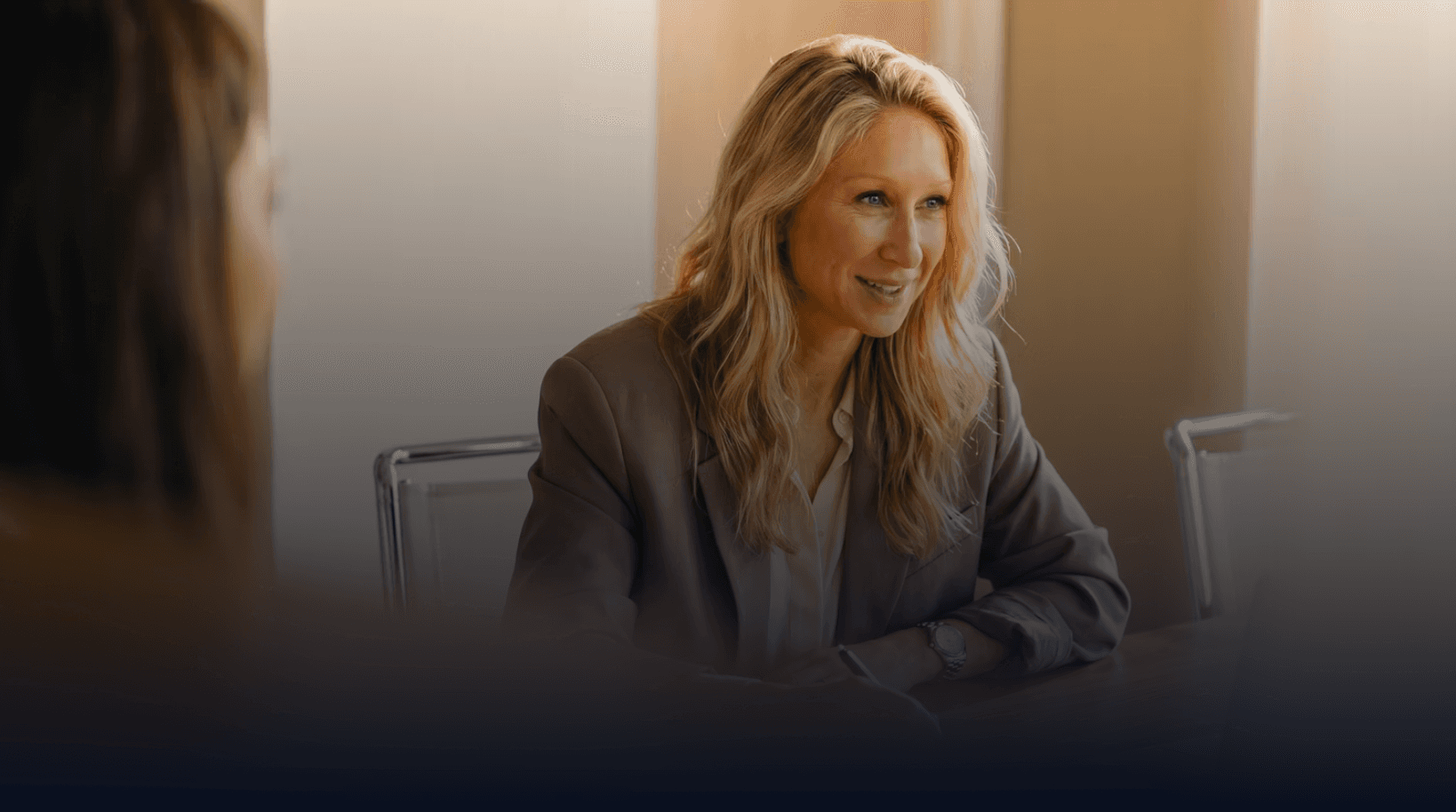 A professional woman in a beige blazer sits at a table, smiling and engaged in conversation in a bright office setting.
