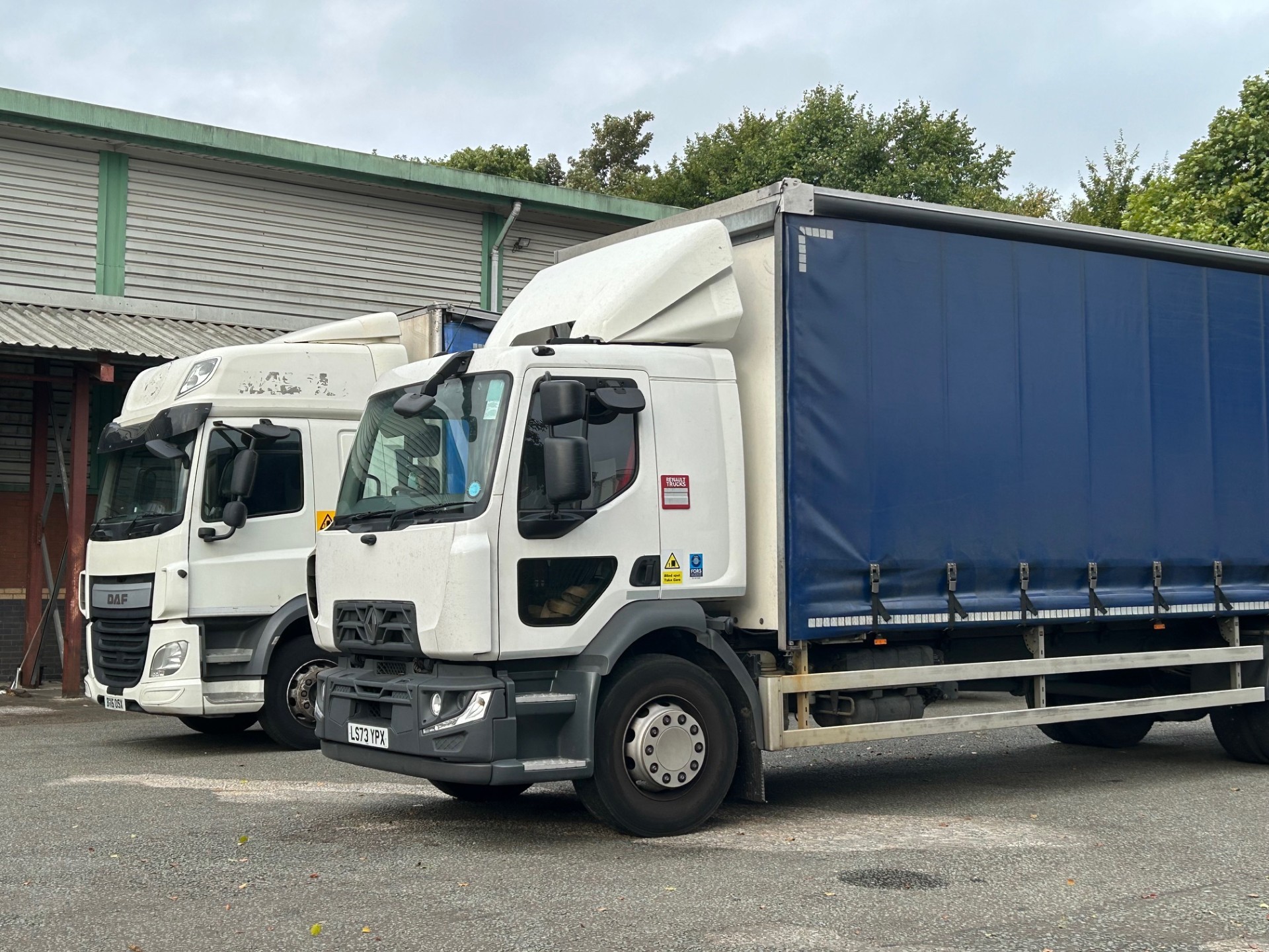 Image of two HGV trucks with blue trailers and while cabs parked outside a warehouse