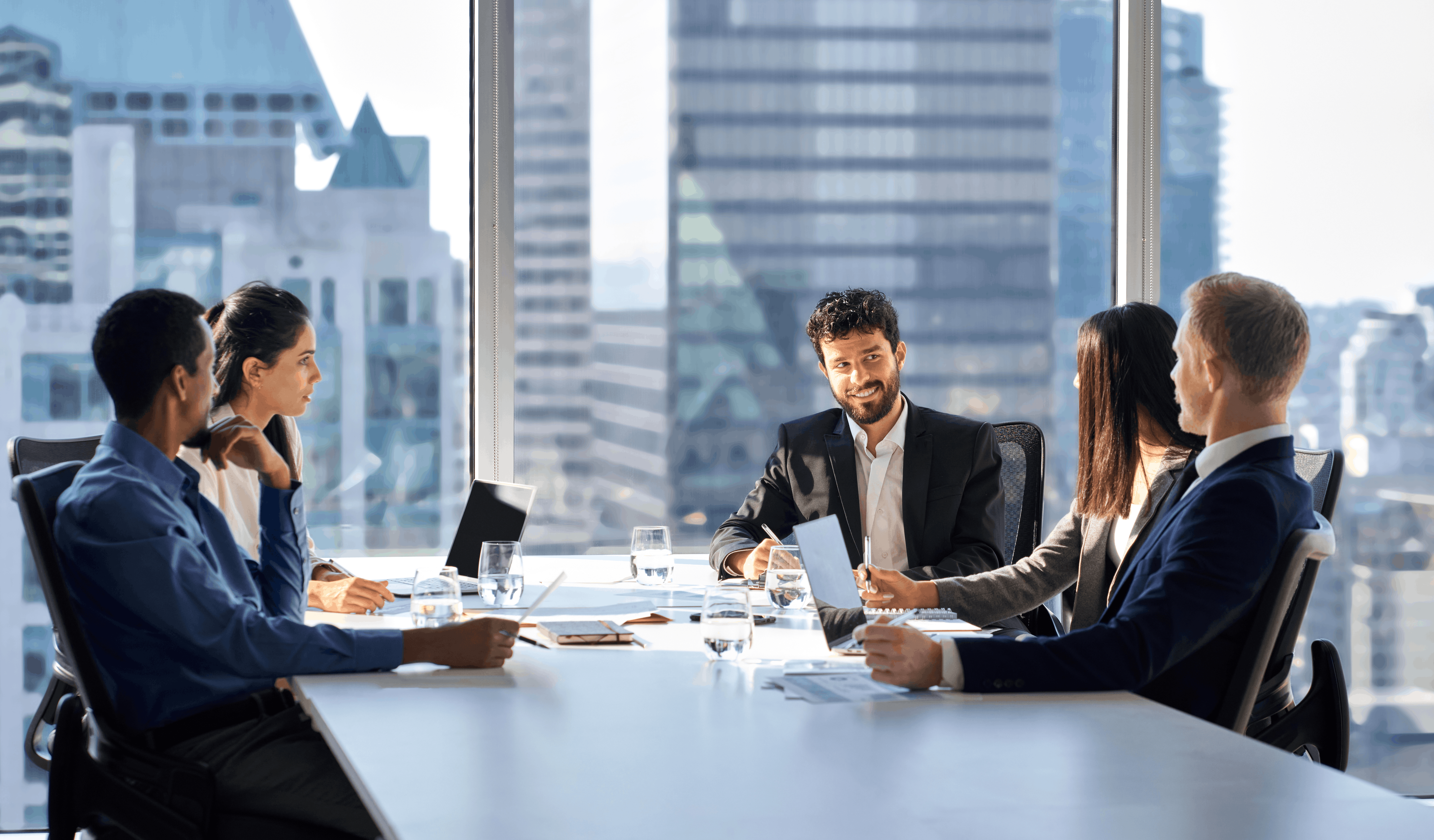 A professional business team seated around a conference table in a bright high-rise meeting room, discussing documents and laptops with a city skyline visible through large windows.
