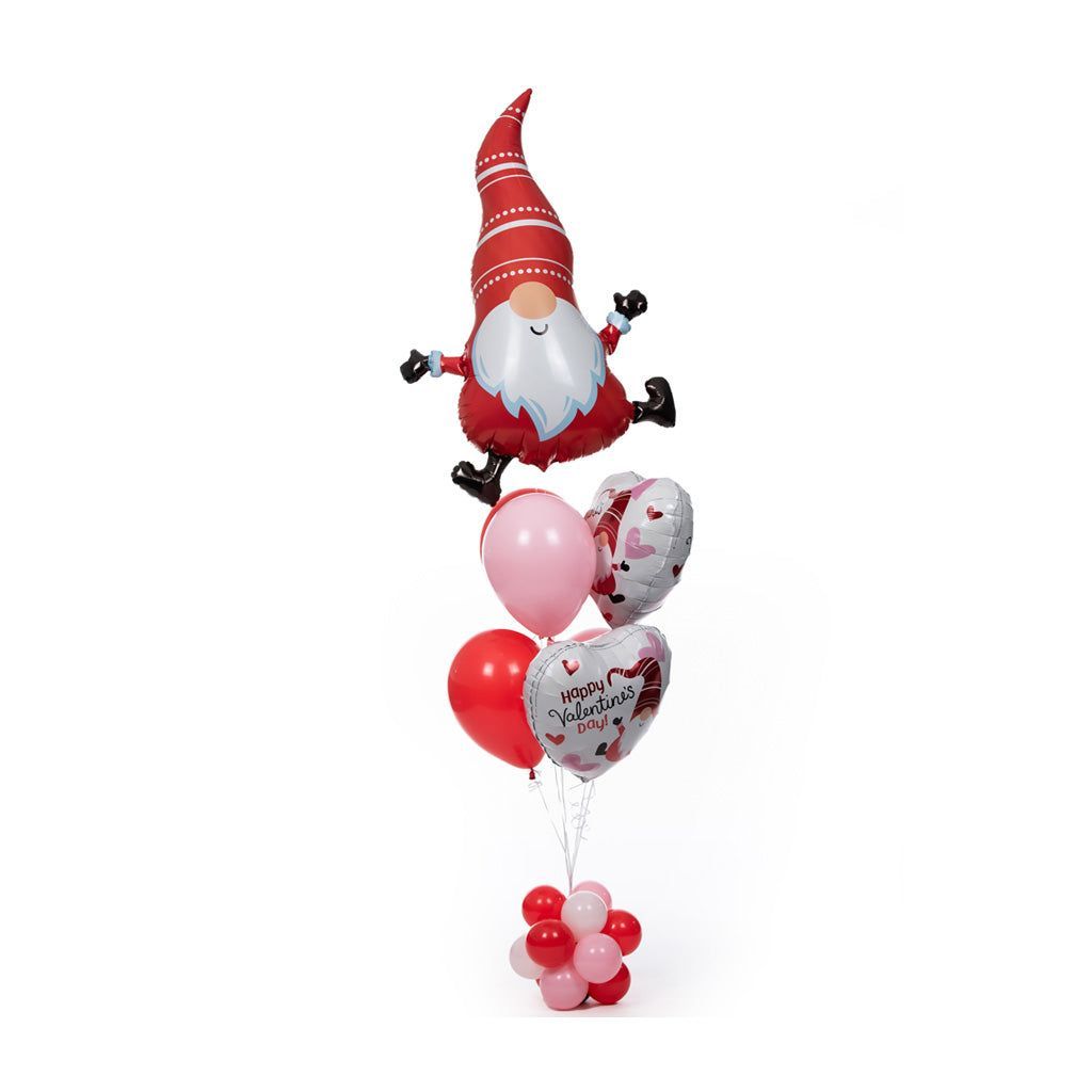 A festive gnome-shaped balloon is perched atop a bouquet of Valentine's Day-themed balloons, featuring red and pink hearts and the message "Happy Valentine's Day," all set against a plain white background.