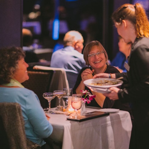 Dalawang babaeng nakangiti sa isang mesa ng restawran habang nagbibigay ng putahe ang isang tagapaglingkod. May mga baso ng alak, kandila, at bulaklak sa mesa.