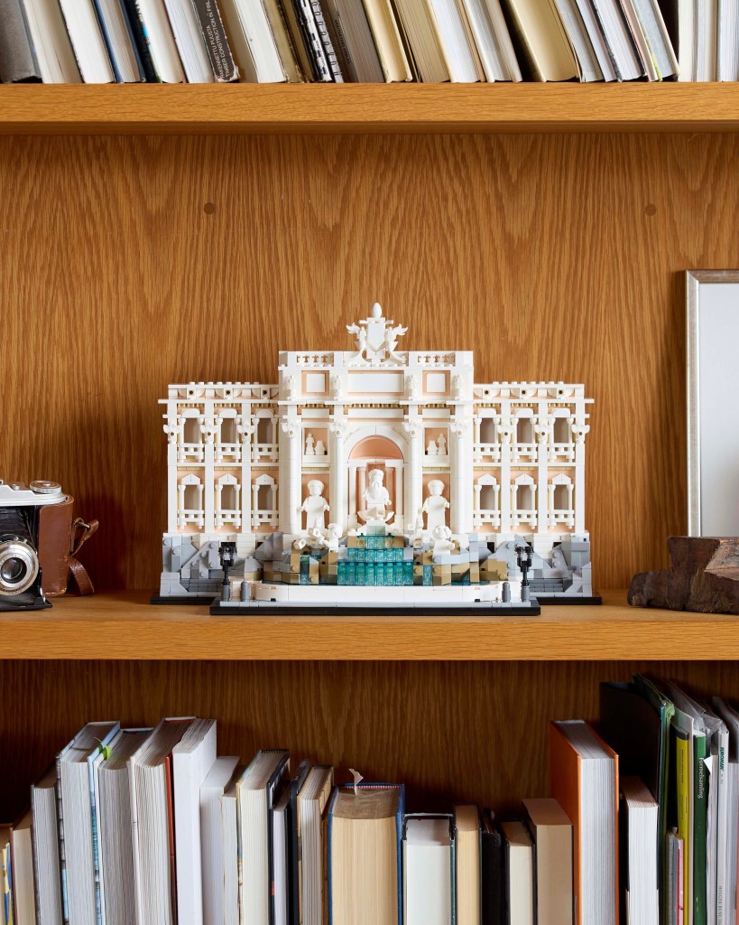 A LEGO model of the Trevi Fountain displayed on a wooden shelf surrounded by books and a vintage camera.