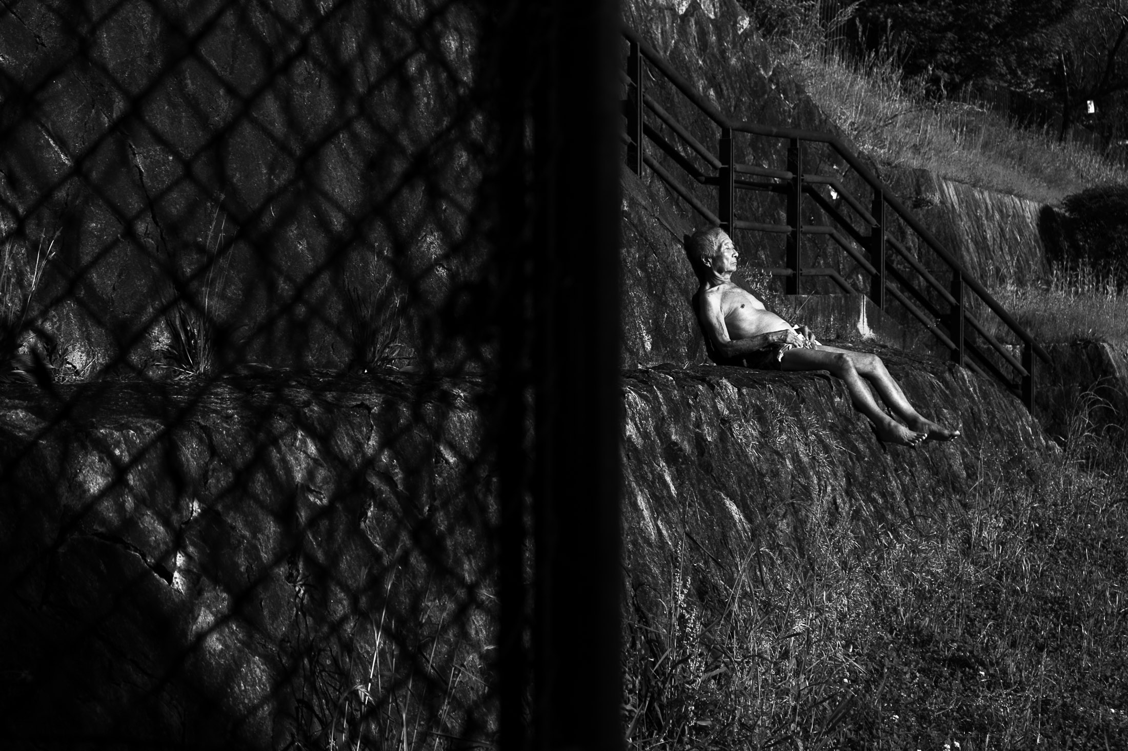 An older man with a bare chest reclines on a sloping stone embankment in the sun while the shadow of a chain-link fence fills the foreground.