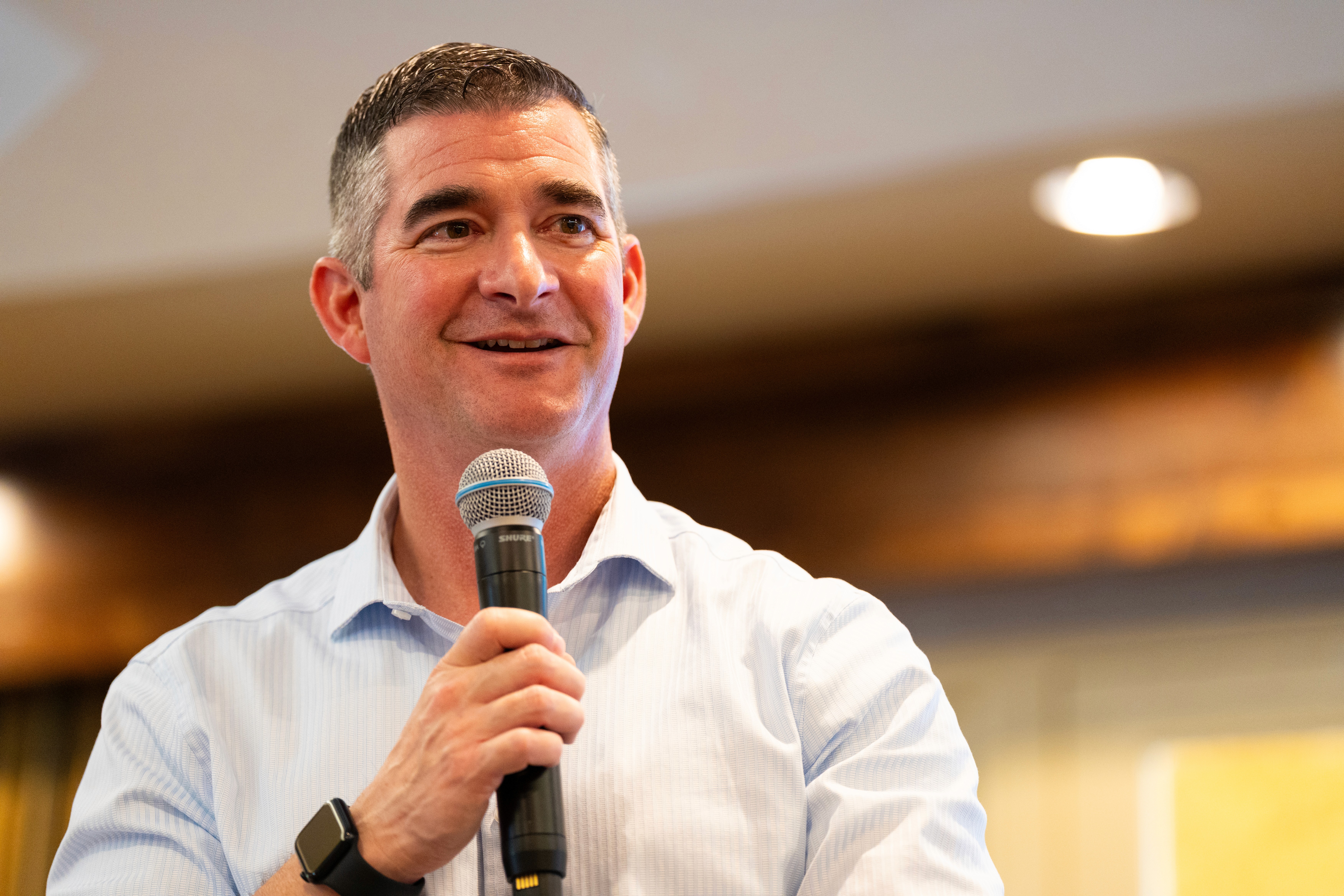 A father carries his daughter on his shoulders as she reaches into an open car trunk. They're by a car and some houses.