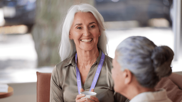 A smiling older woman with white hair wearing a lanyard, engaged in conversation with another person in a professional setting