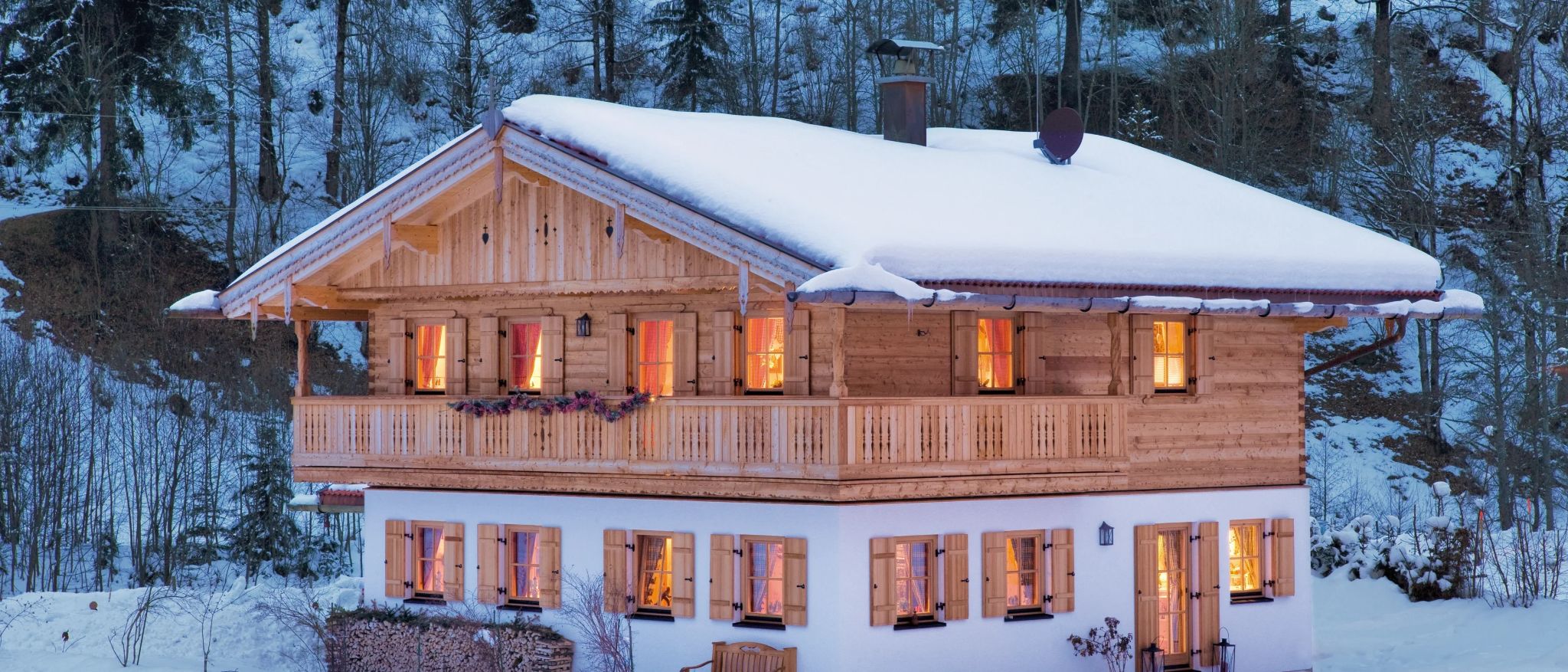 Holzhaus im Schnee mit leuchtenden Fenstern, Balkon mit Holzschnitzereien und Holzstapel im Vordergrund.