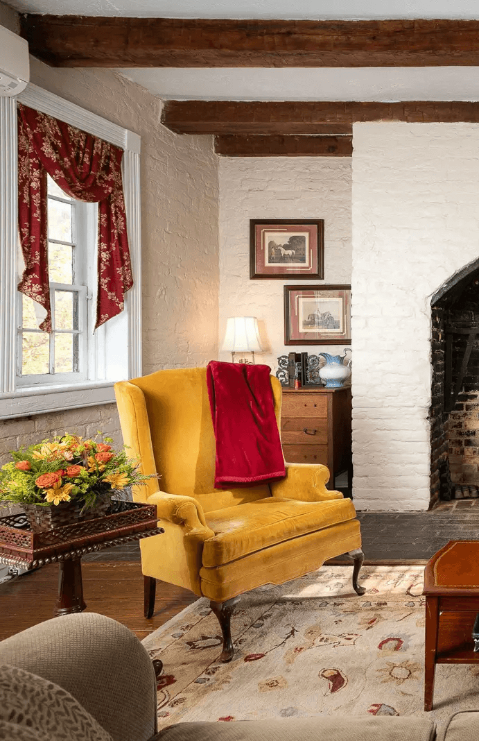 A cozy living room features a mustard yellow wingback chair draped with a red throw, positioned near a rustic brick fireplace, accented by wooden beams and decorated with floral curtains and framed artwork.