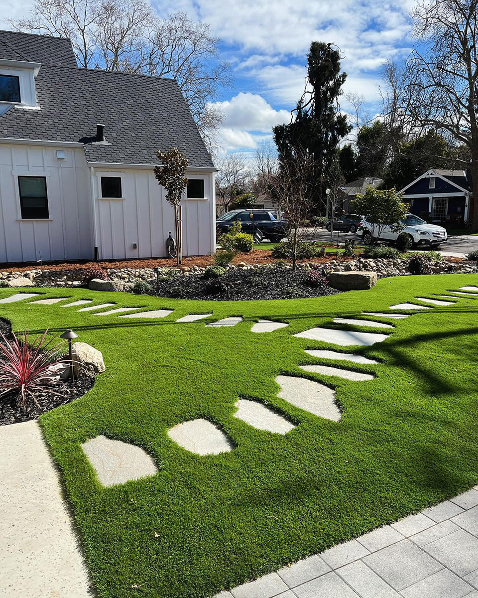 A picture of a lush green artificial lawn by AGL Grass with a winding stone path leading up to a charming house in the background.