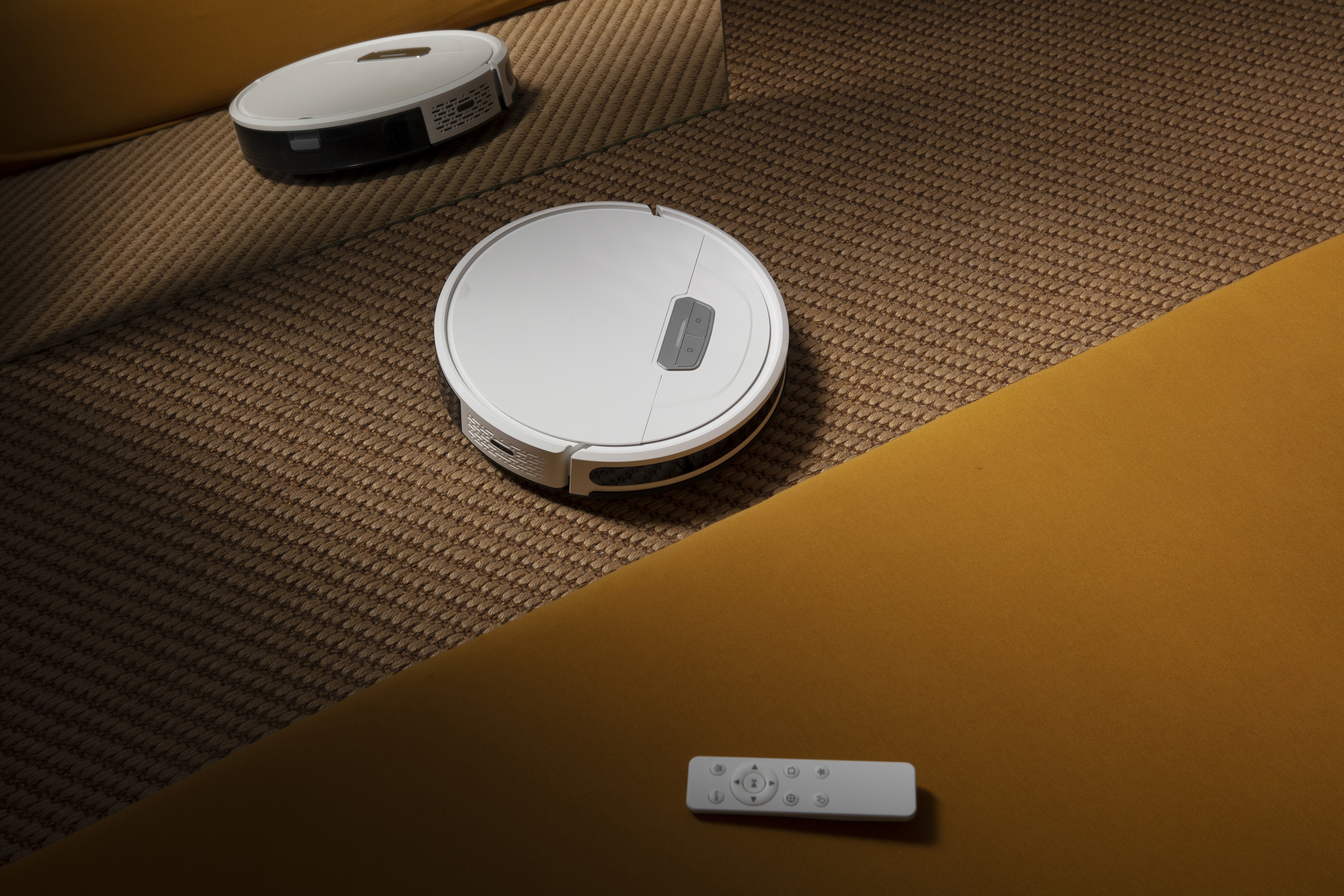 White robot vacuum cleaner on carpeted floor with remote control nearby.