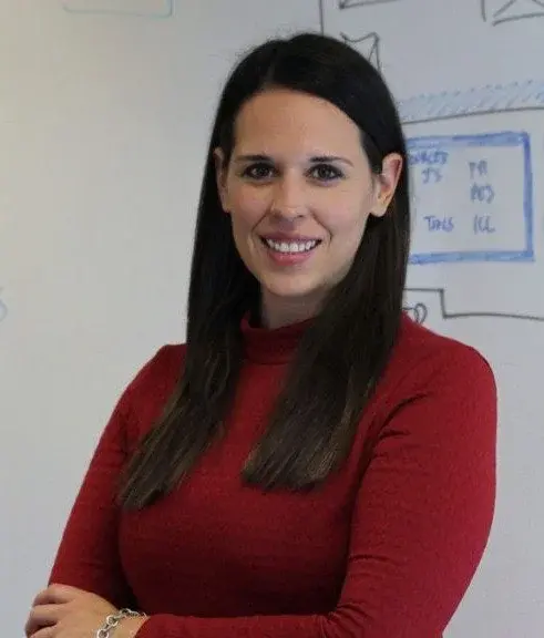 A person with long dark hair, wearing a red sweater, stands smiling with arms crossed in front of a whiteboard covered in sketches and notes in an office setting.