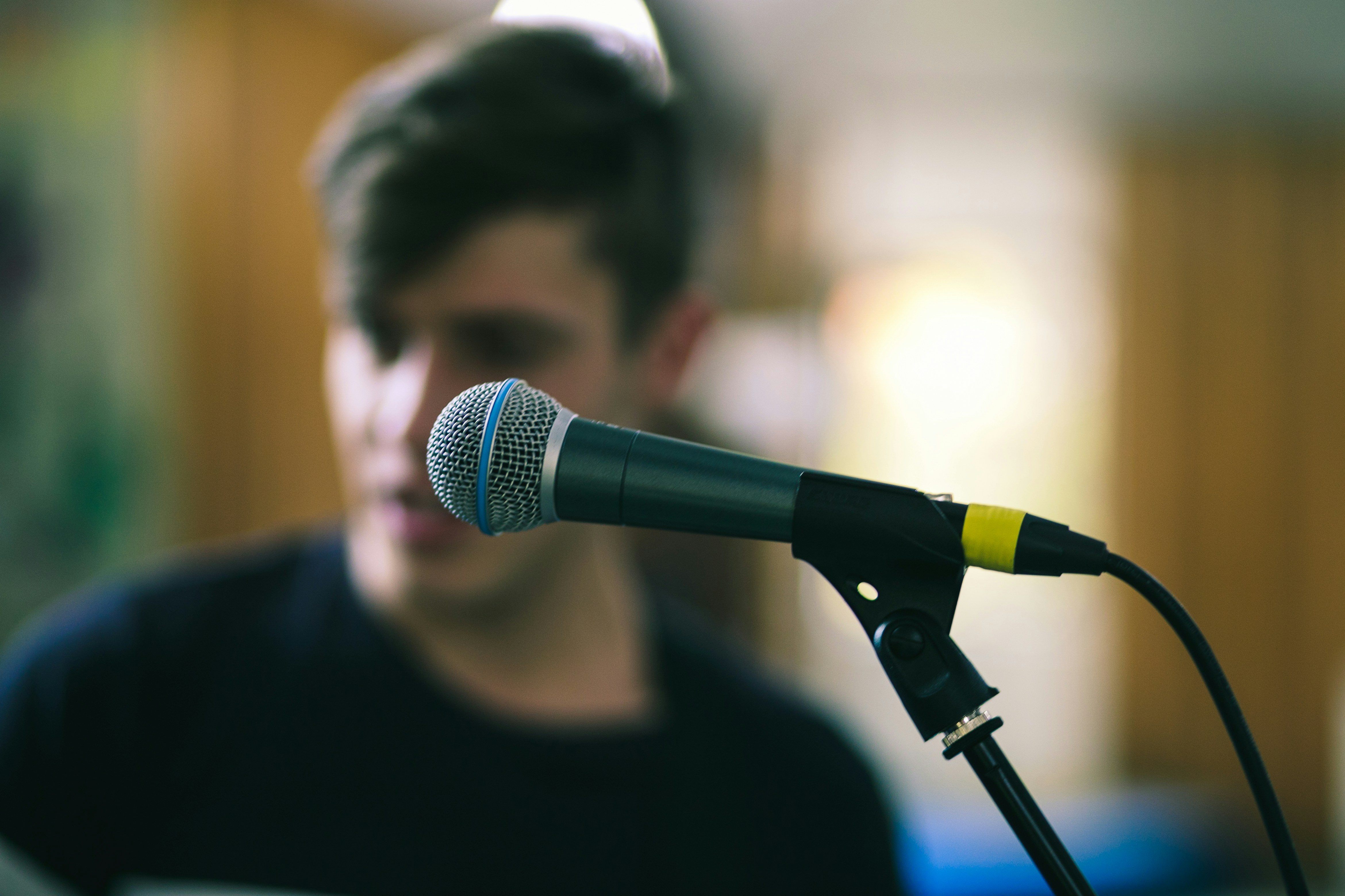 A microphone setup for vocal performance and a singer out of focus in background. Photo by James Coleman on Unsplash