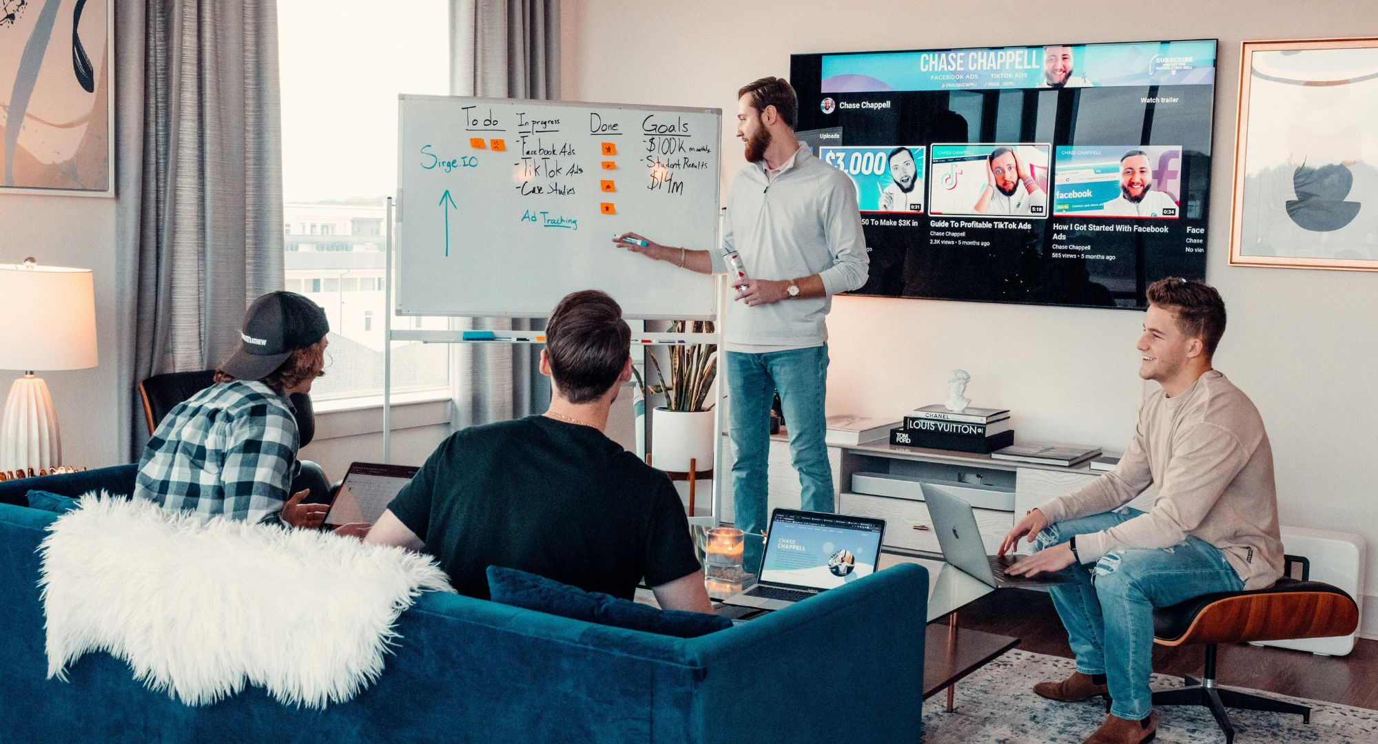 quatro pessoas numa sala em uma reunião de planejamento, três estão sentadas e uma está em pé explicando junto a um quadro