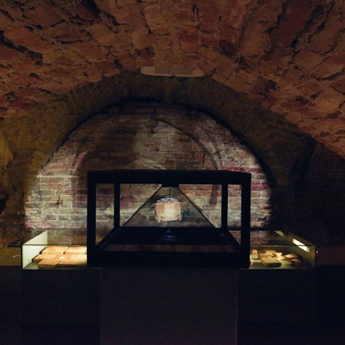Dimly lit brick-walled room with an illuminated display case holding an artifact, flanked by illuminated shelves.