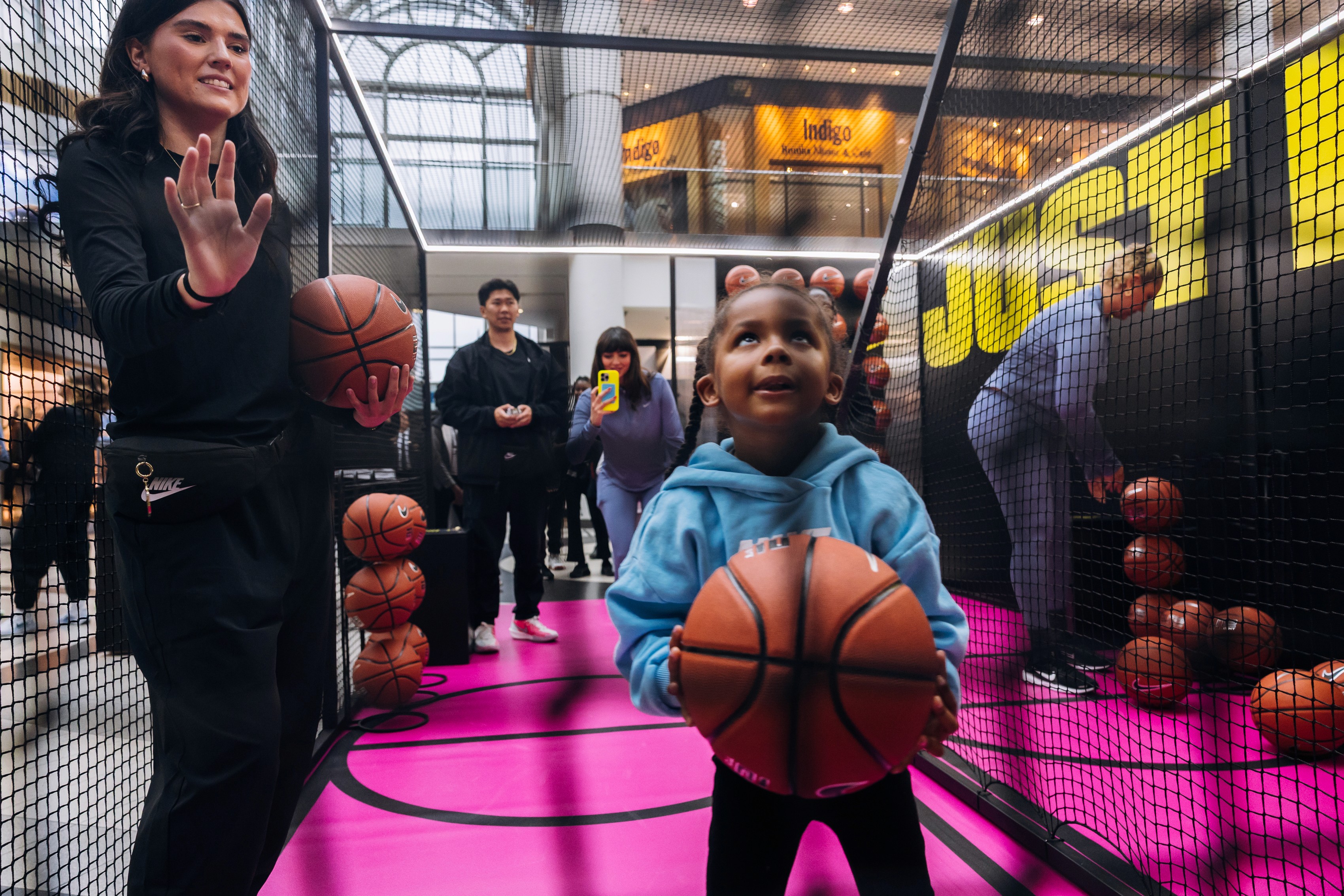 A young child in a blue hoodie holds a basketball, looking up with excitement in an indoor basketball setup. An adult nearby guides them. Bright, playful atmosphere.