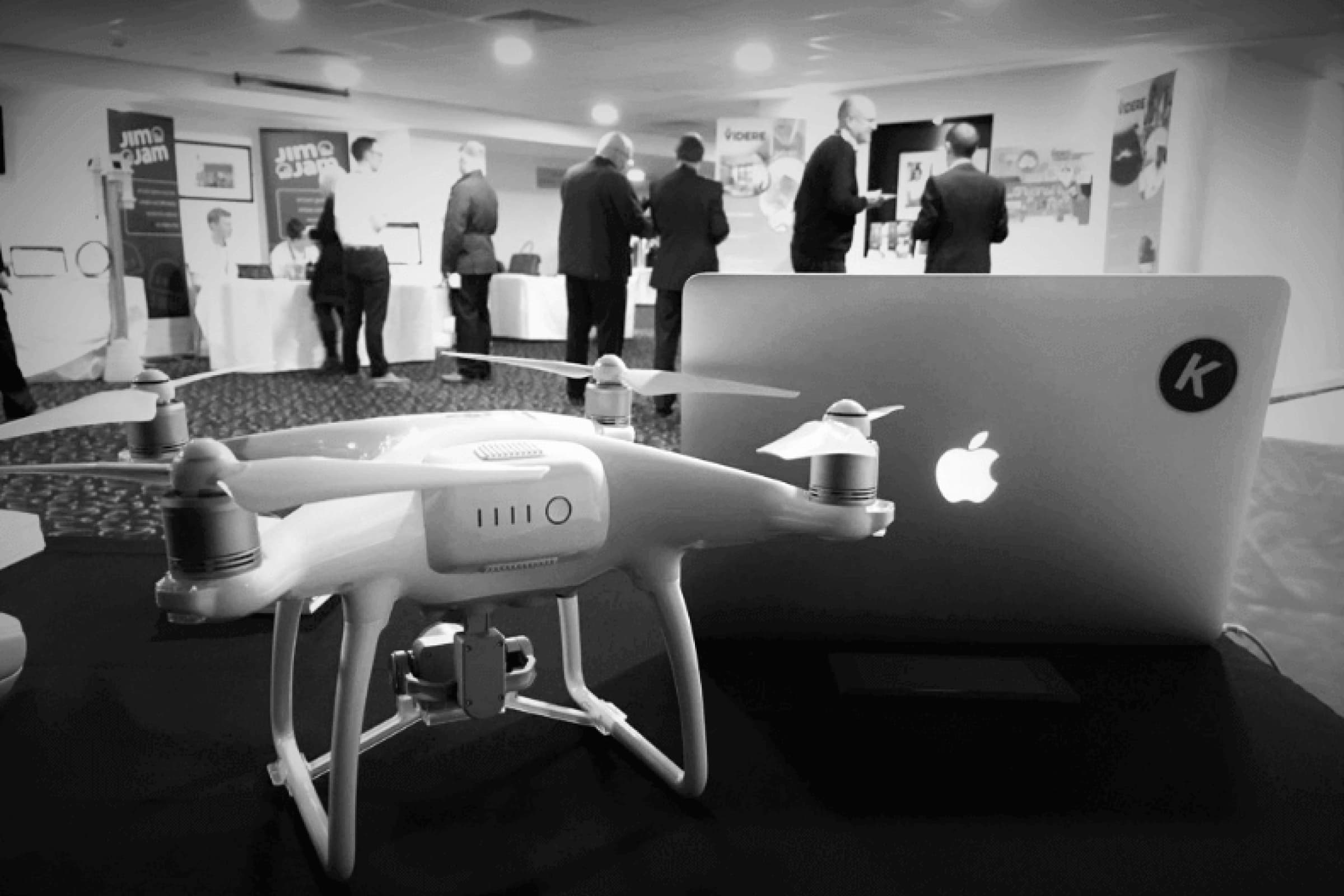 Venturefest conference photo in black and white, with a drone and laptop in the front, with some people chatting in the background