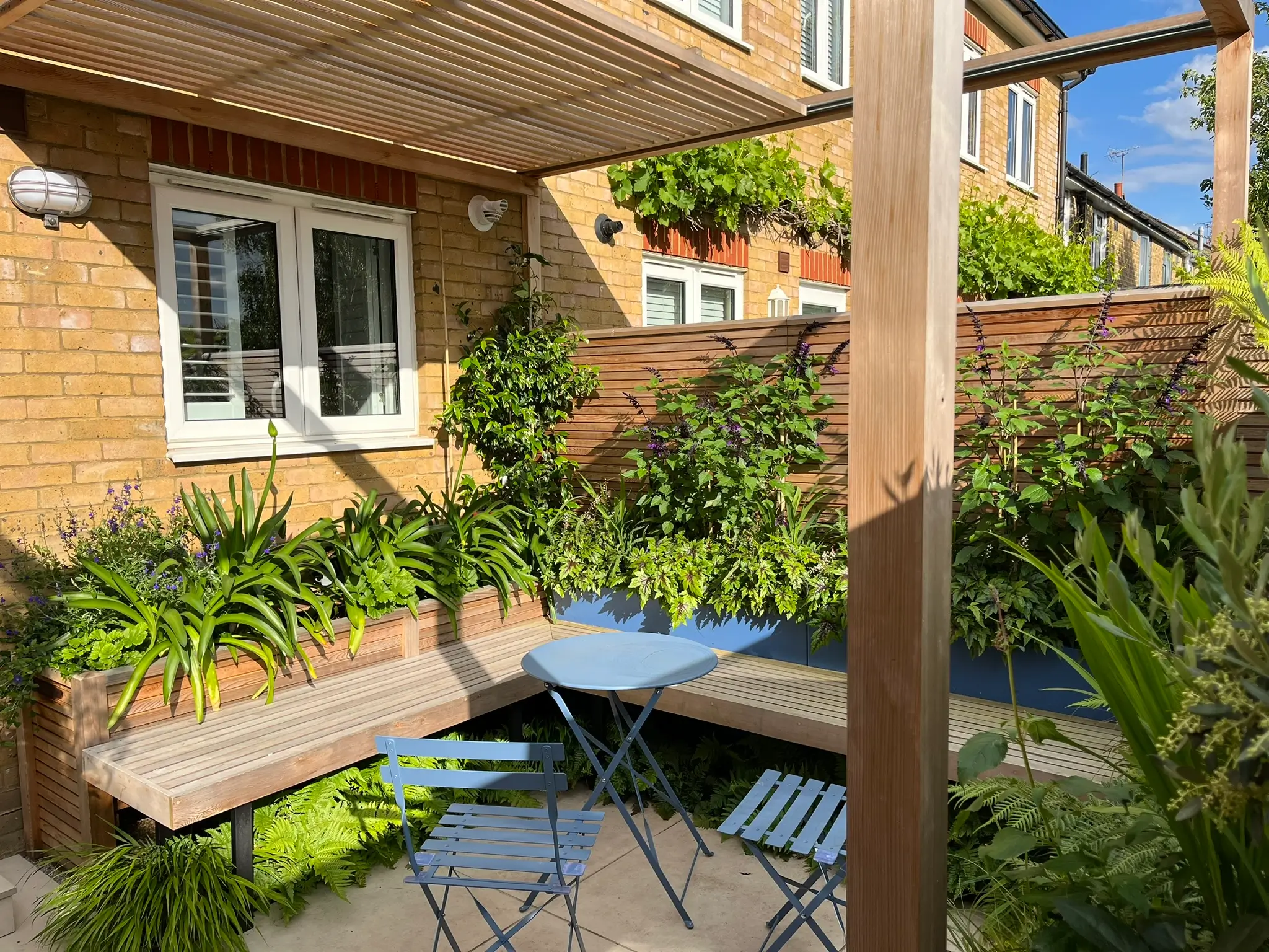 A cozy outdoor garden scene with a small table and chairs, surrounded by lush greenery and a sunny atmosphere.