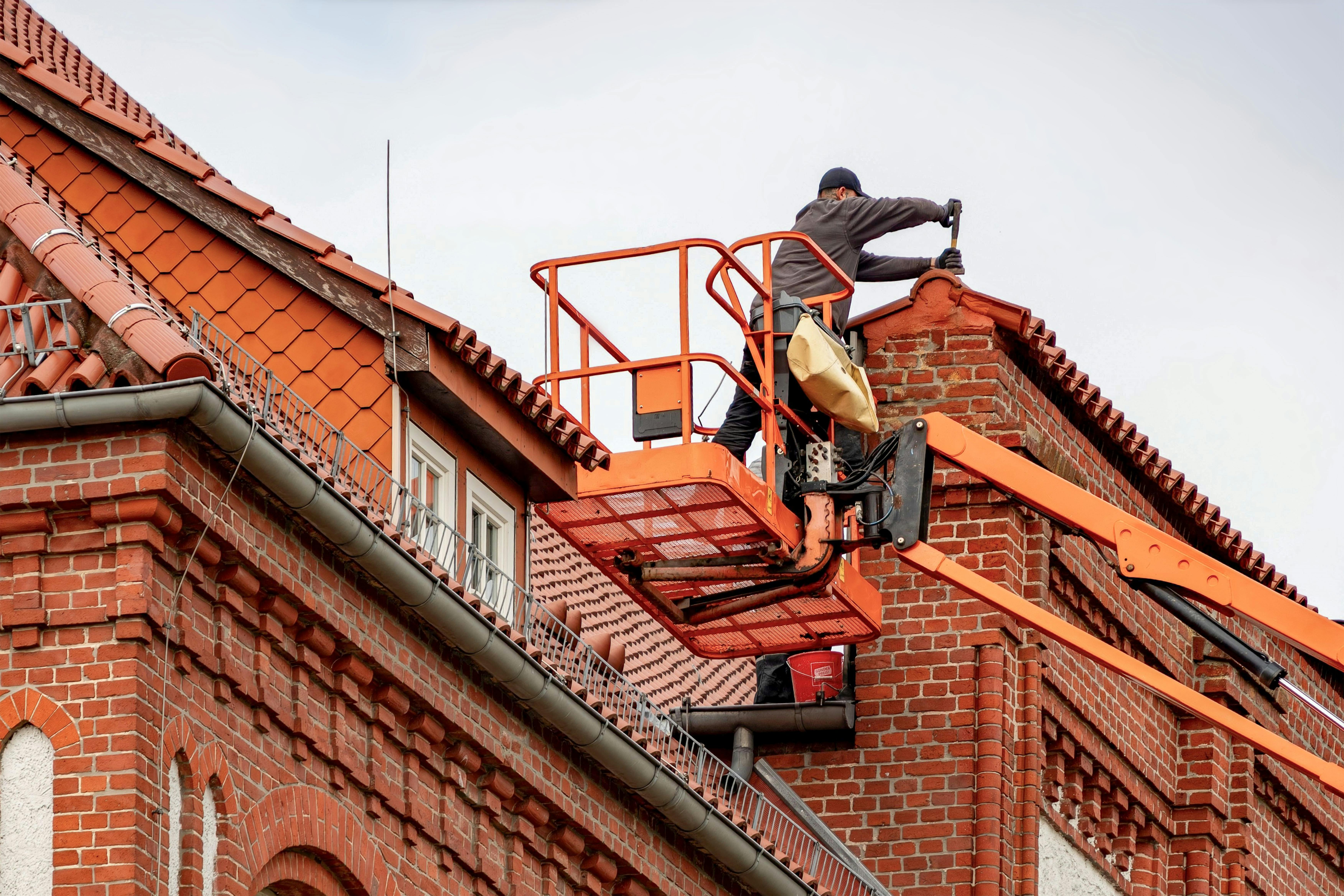 Technische vakman voert onderhoudswerkzaamheden uit op een dak met behulp van een hoogwerker bij een bakstenen gebouw, gericht op veilig werken op hoogte in de bouw en techniek.
