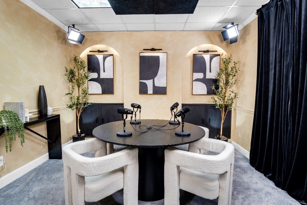 Book Studio C “Studio C with a round table and four white upholstered chairs, set against a beige wall with abstract art, black curtains, and visible studio lighting equipment for a creative workspace feel.”