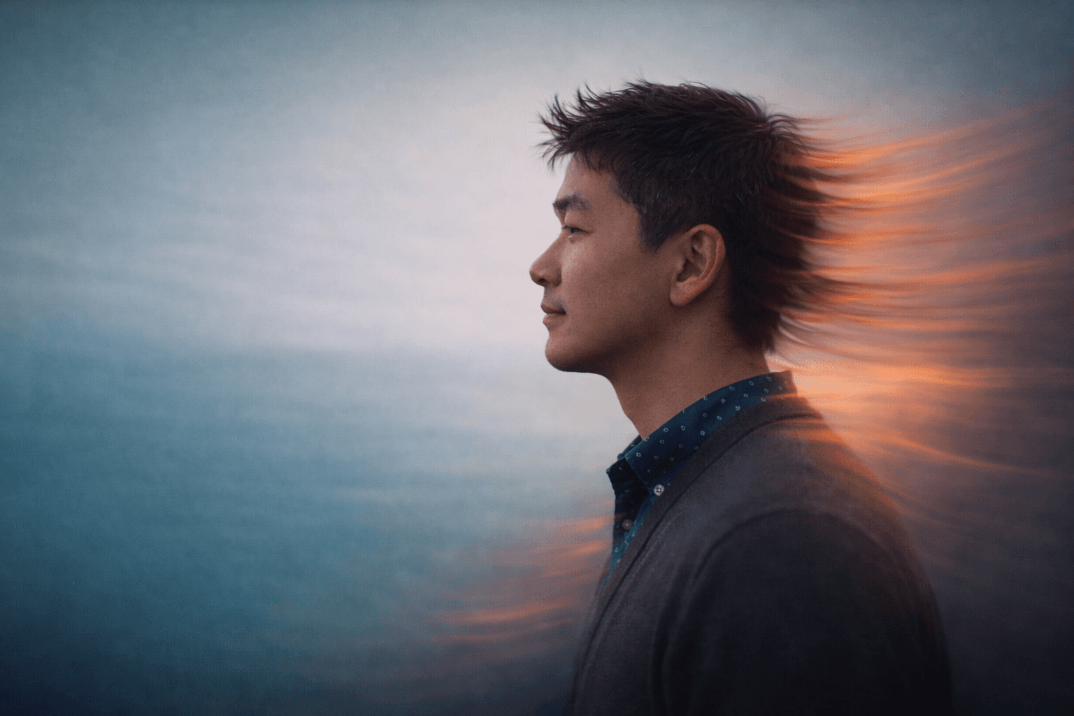 Side-profile portrait of a person facing left in a blazer and patterned shirt, set against a soft blue-gray gradient background with warm orange light streaks trailing behind their head and shoulder.