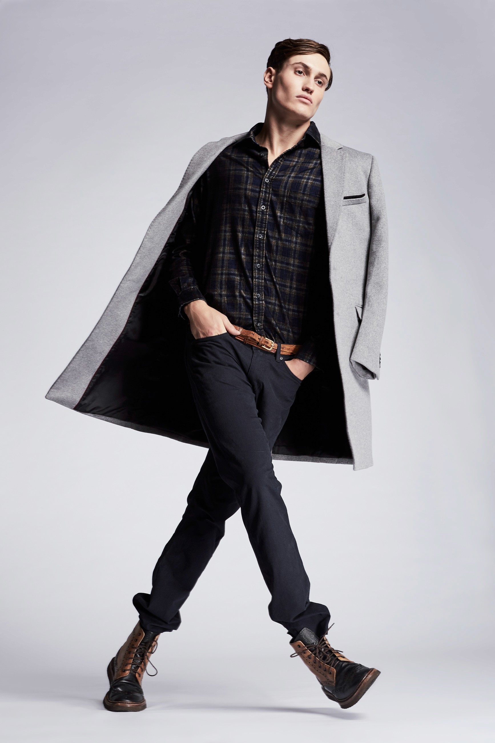 A male model off-balance and mid-stride on a minimal white background, hands in dark blue jean pockets, and wearing a dark shirt with a light grey duffel jacket around his shoulders and brown leather boots.