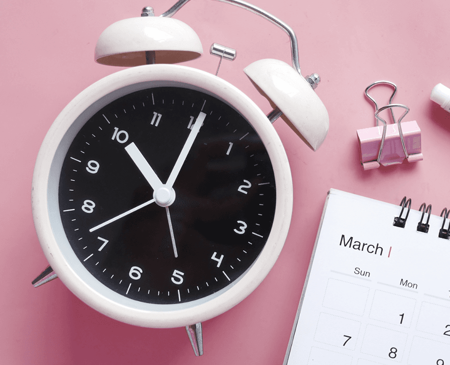 A white vintage-style alarm clock with black face showing approximately 11:58, positioned on a soft pink background alongside office supplies including a pink binder clip and a partial view of a March calendar.