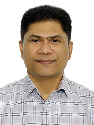 Man with a bald head, beard, and glasses, wearing a blue suit and white shirt, stands confidently against a white, ornate wall. Professional and composed.