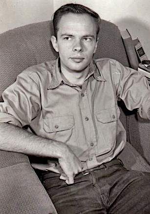 Black and white image. A man sits in an armchair, wearing a button-up shirt and jeans. He has short hair and rests one arm on the chair. The setting appears to be indoors, with a telephone visible in the background.