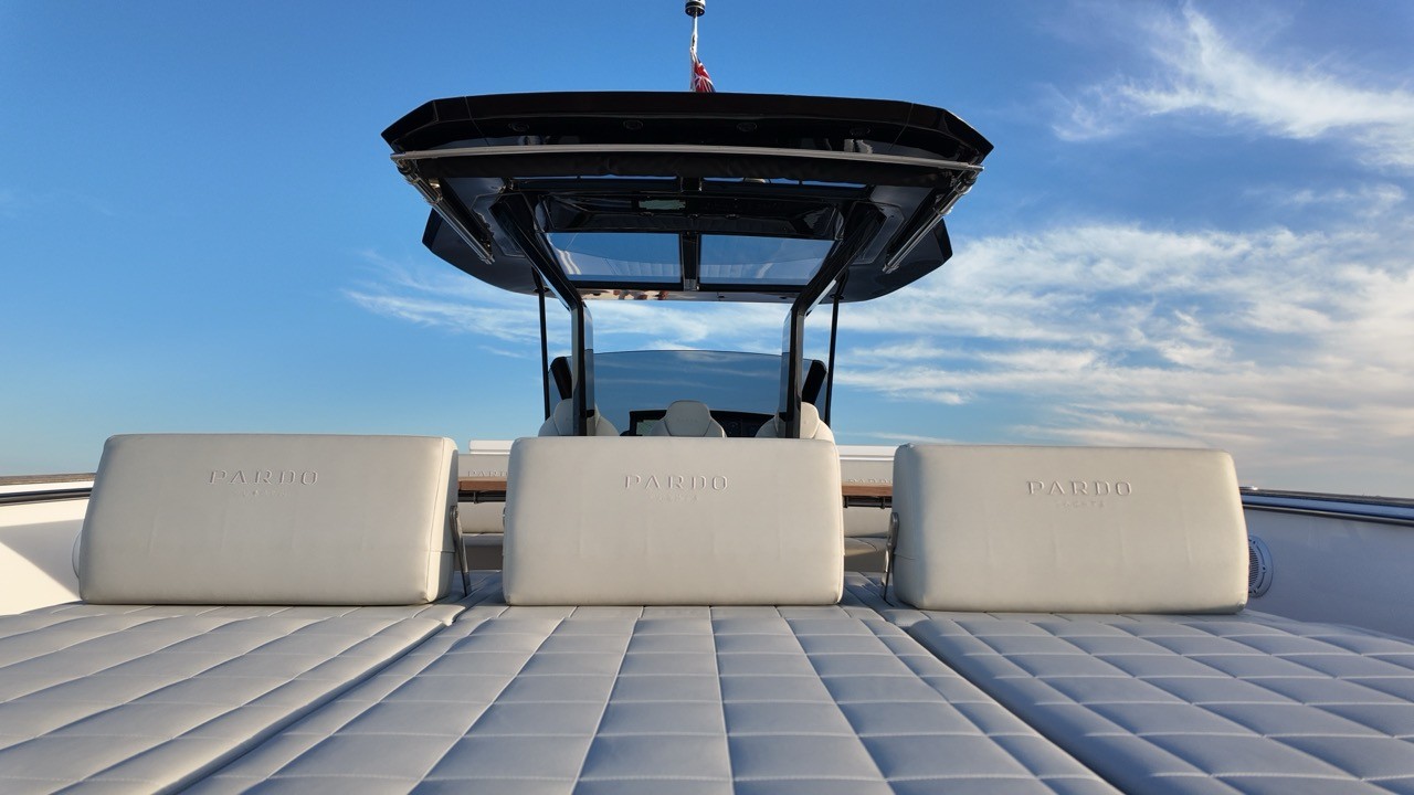 View from the Pardo 43 foredeck sunpad looking aft toward the helm station, showing the quilted grey sunpad cushions, three Pardo-branded white leather backrests with embossed logo, black T-top hardtop with glass roof panel open to the sky, and helm seating visible beyond against a bright blue sky