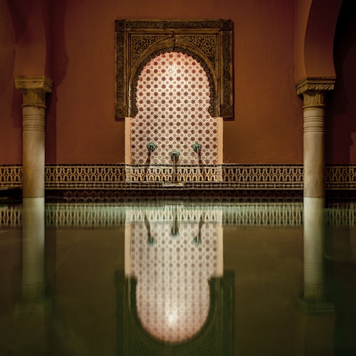 華麗的室內游泳池，配有柱子和複雜的瓷磚圖案，拱形噴泉有三個水口，安靜的水面反射著裝飾。