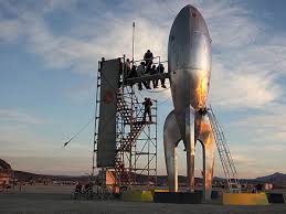 A large, silver, retro-style rocket sculpture with fins stands in a desert-like landscape. Next to it is a tall scaffold tower with stairs, where a group of people is gathered on a platform overlooking the ground. The scene is set under a dramatic twilight or sunset sky.
