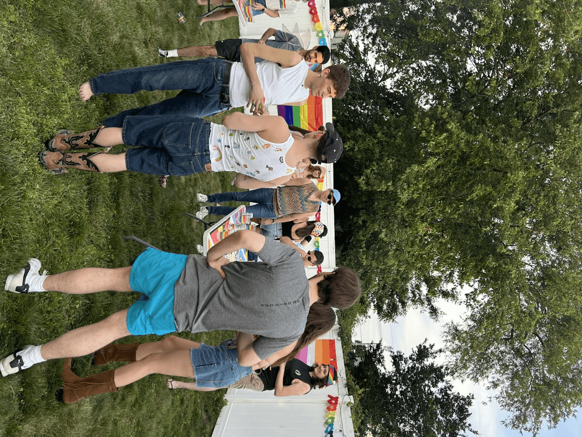 A group of people gather outdoors, enjoying a sunny day in front of colorful rainbow flags.