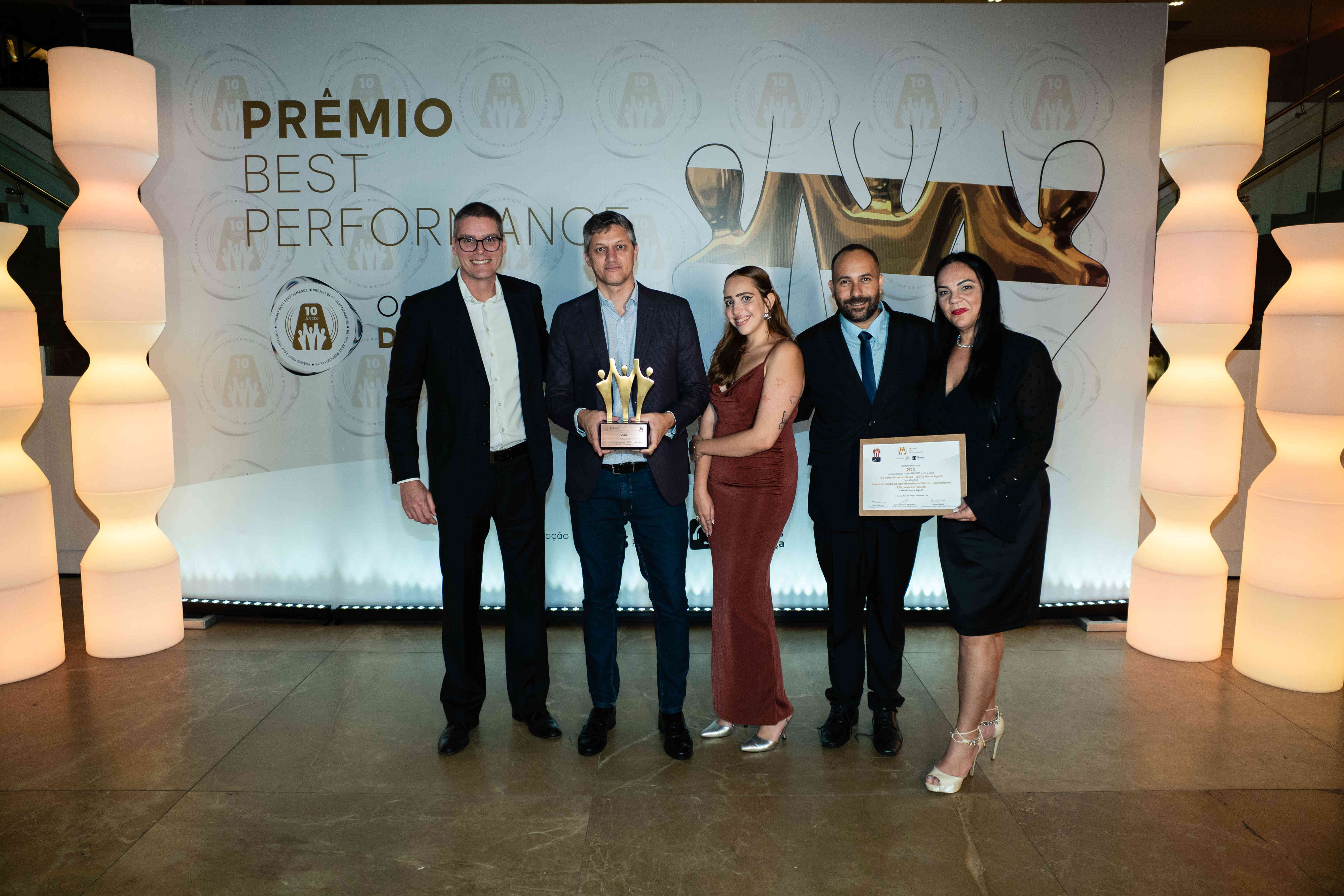 Executivos da 2CX posando com troféu bronze e certificado no palco do Prêmio Best Performance 2025
