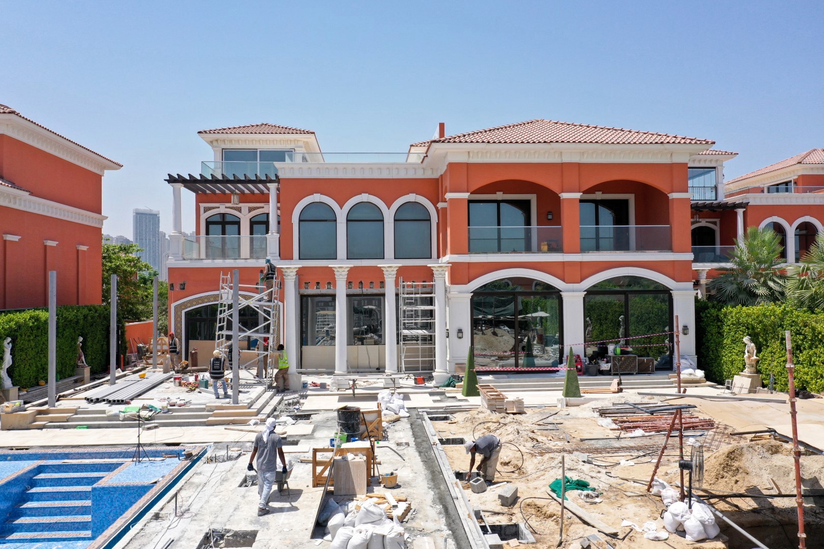 A luxurious Mediterranean-style villa with arched windows and terra-cotta roof tiles is undergoing significant construction and landscaping work in the backyard, including a partially built swimming pool and various building materials scattered around, framed by neatly trimmed hedges and distant city skyline.