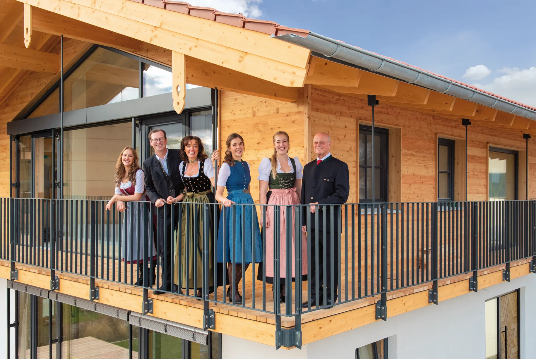 Eine Familie in Tracht steht auf einem Balkon vor einem Holzhaus.