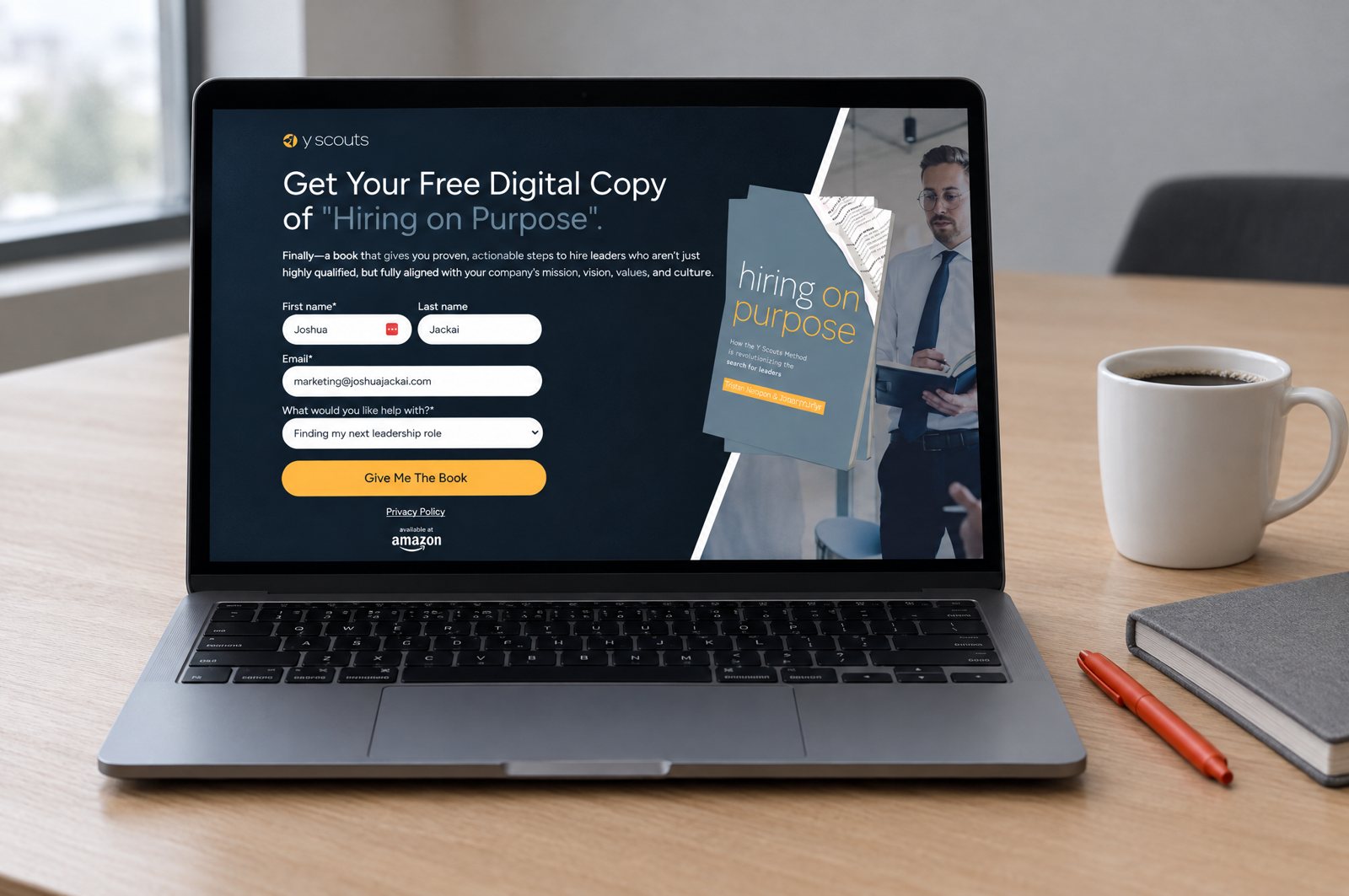 A laptop on a meeting-room table showing the real Y Scouts landing page built in HubSpot by Demand Designer: 'Get Your Free Digital Copy of Hiring on Purpose' with a short form and a 'Give Me The Book' button; a red-orange pen and a mug beside it