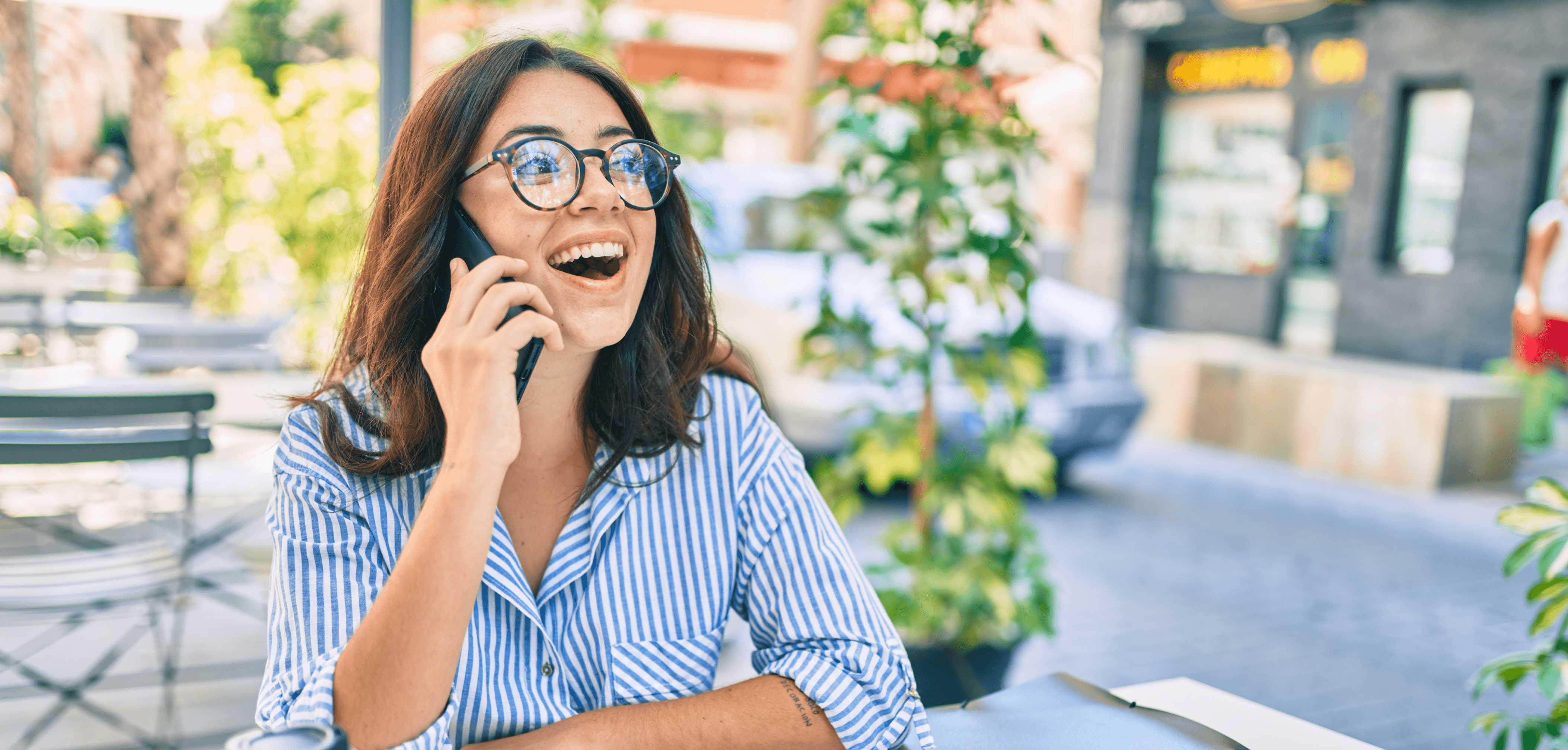 Smiling person wearing glasses, talking on a smartphone while sitting at an outdoor café.