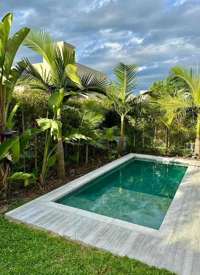 Espectacular piscina de hormigón con agua verde esmeralda, enmarcada por un borde atérmico claro y un hermoso parque con palmeras.
