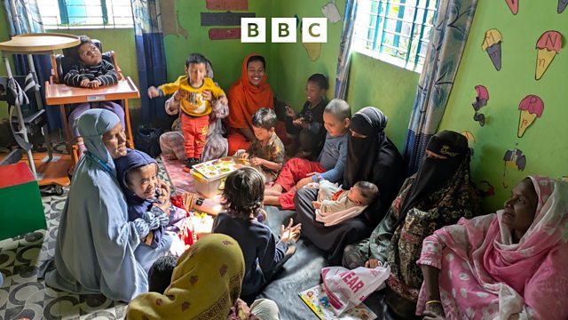 A group of women and children gathered together, sitting on a floor mat in a colorful room with green walls decorated with cutouts. One child is seated in a supportive wooden chair. The BBC logo is overlaid at the top center.