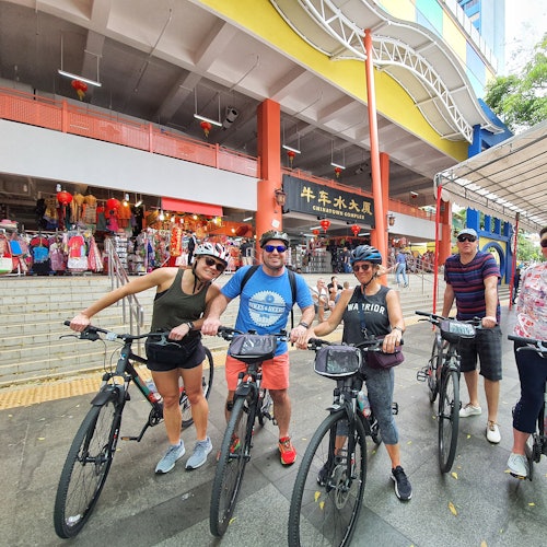 นักปั่นจักรยานสี่คนโพสท่าหน้าตึกพาณิชย์สีสันสดใสที่มีร้านค้านานาชนิดและป้ายโฆษณา พวกเขายิ้มแย้มและสวมหมวกกันน็อค