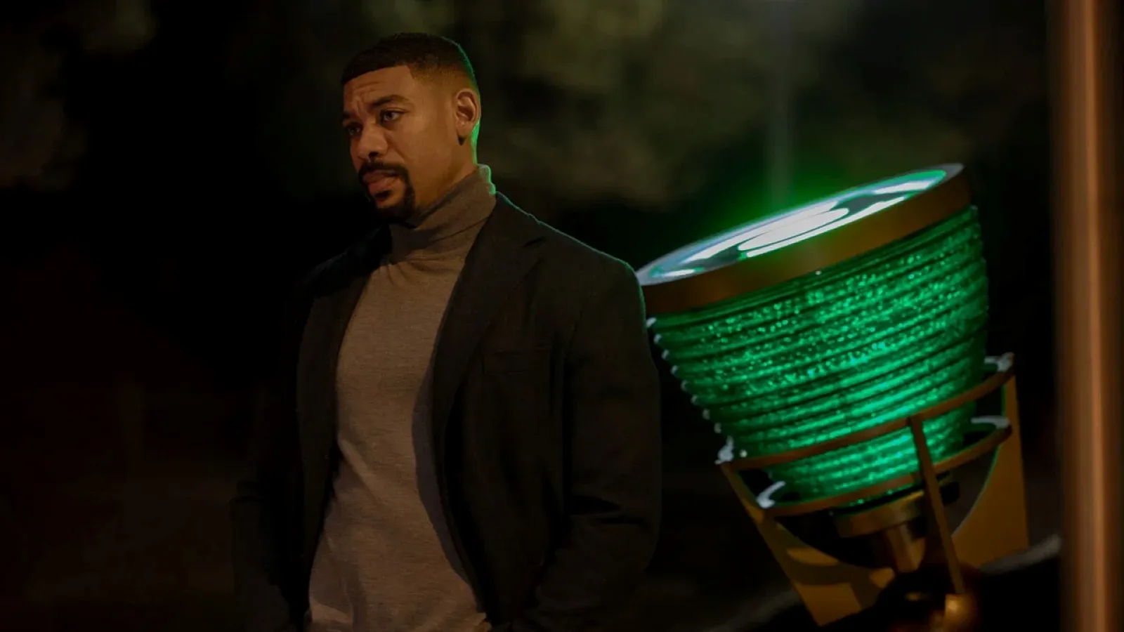 A man in a dark suit and grey turtleneck stands outdoors at night next to a large, glowing green lamp, thoughtfully gazing into the distance.