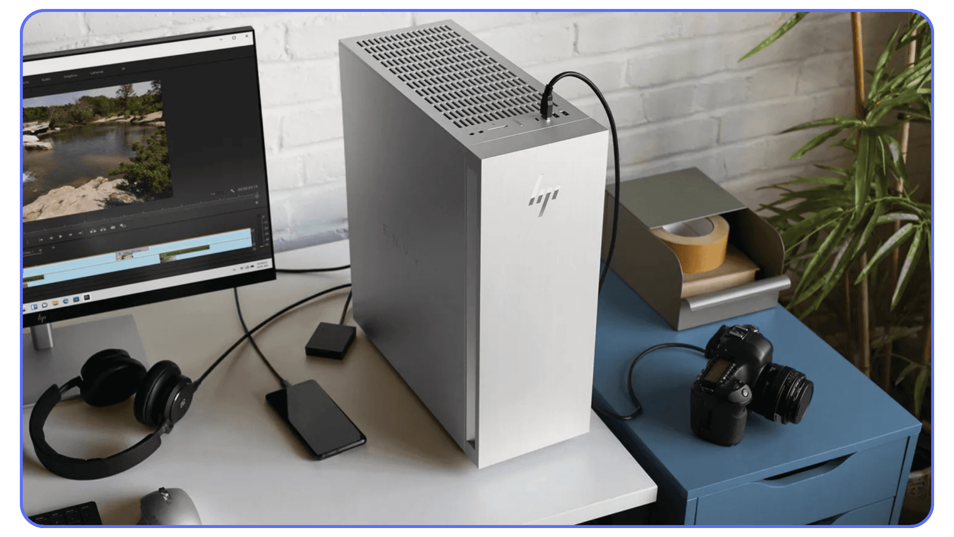A silver HP Envy desktop next to a monitor displaying a video editing screen, with headphones, a camera, and other accessories on the desk.