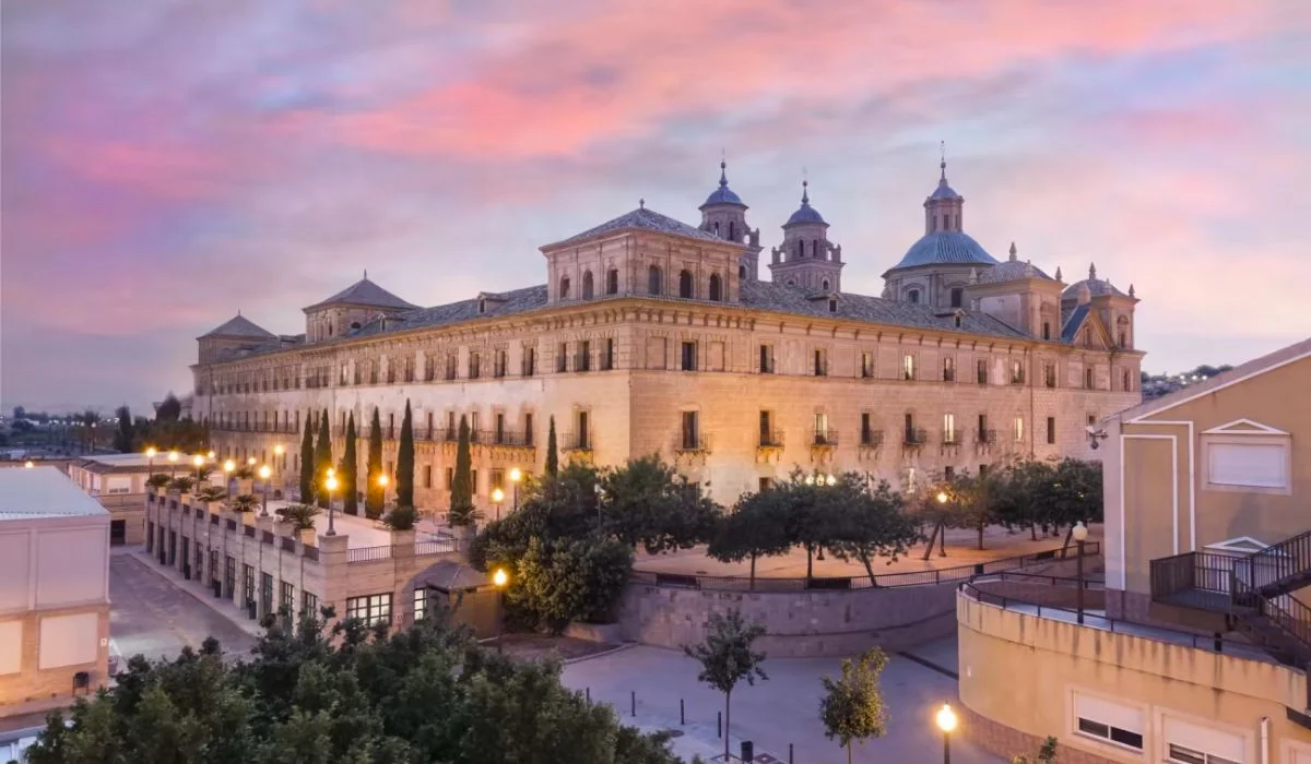 The historic building of UCAM University stands majestically at sunset, illuminated by warm lights and surrounded by lush trees, creating a picturesque architectural scene.