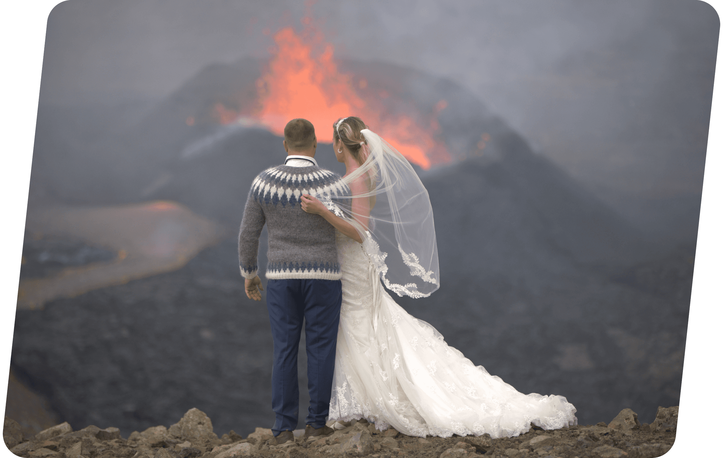 A bride in a white wedding dress and veil stands beside a groom wearing a patterned sweater and blue trousers, both facing an erupting volcano with glowing lava in the background.