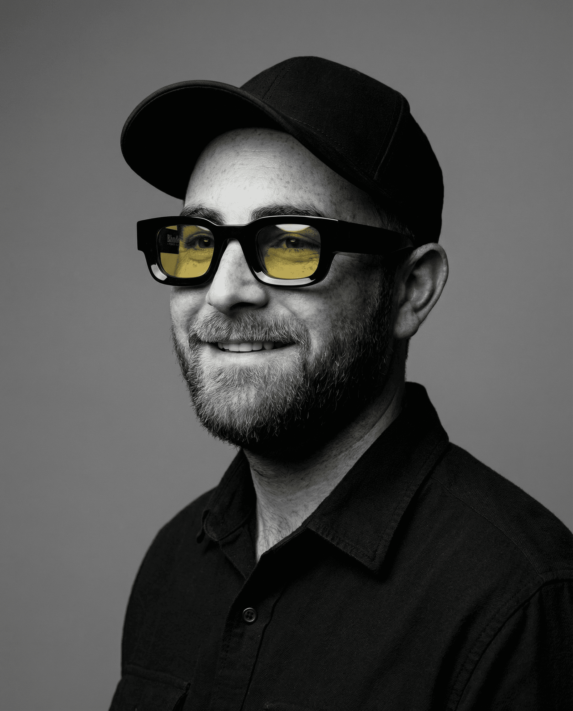 Smiling young man with curly hair, wearing a dark hoodie, against a softly lit background.