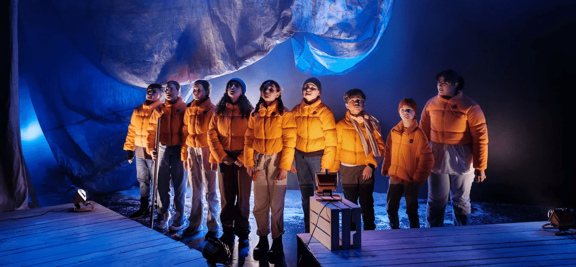 Ten performers stand side by side in orange puffer jackets, gazing upward in awe. They are lit by a cool blue glow beneath a large, rippling tarp that evokes a frozen or icy landscape.