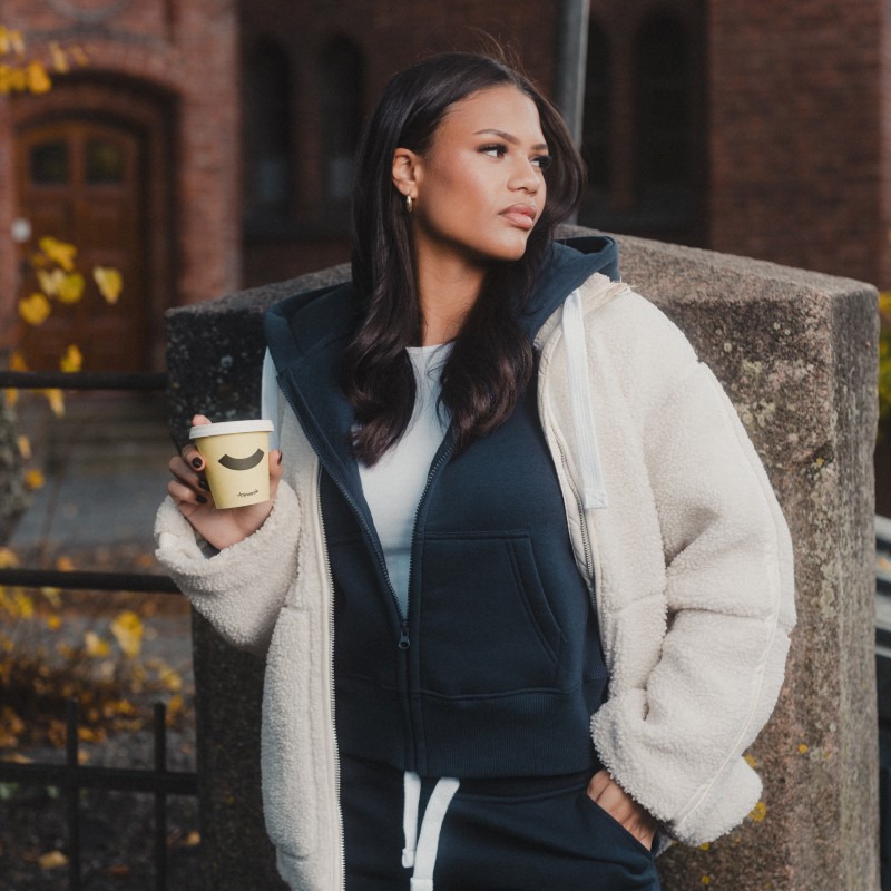 Young woman wearing a navy ziphood and fluffy white jacket holding a coffee-cup