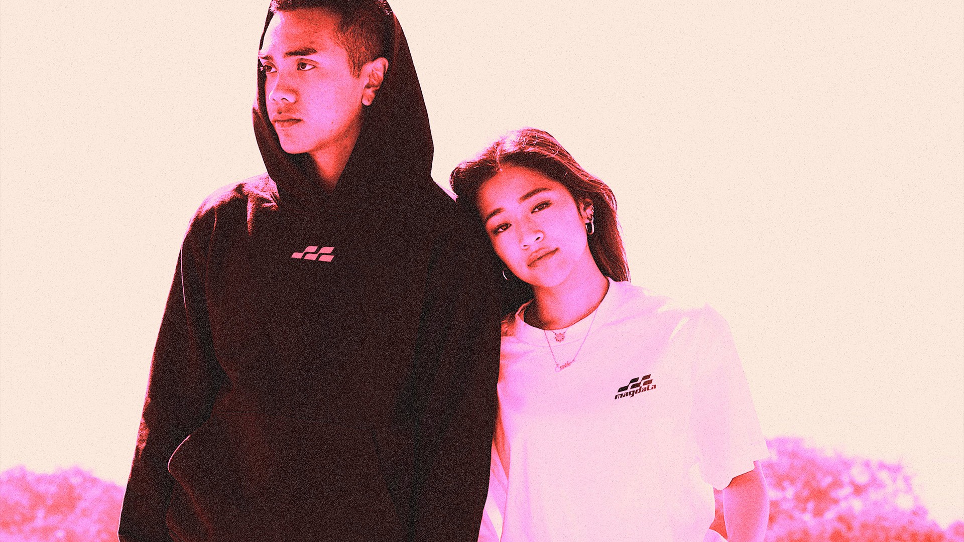 Two young people wearing black hoodies standing together against a pale pink background.