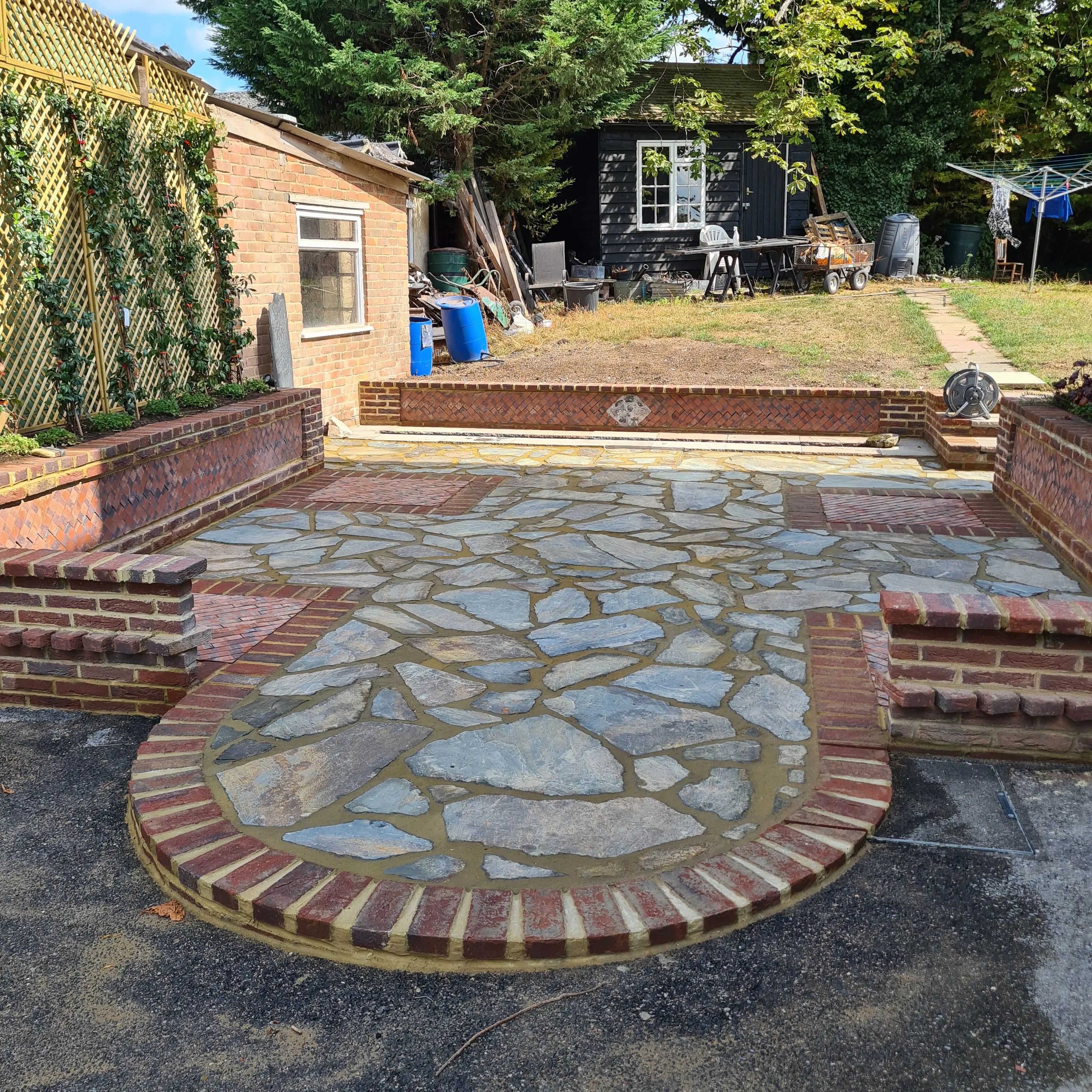 A stone mosaic table sits on a patio, surrounded by brick seating and lush greenery in the background.